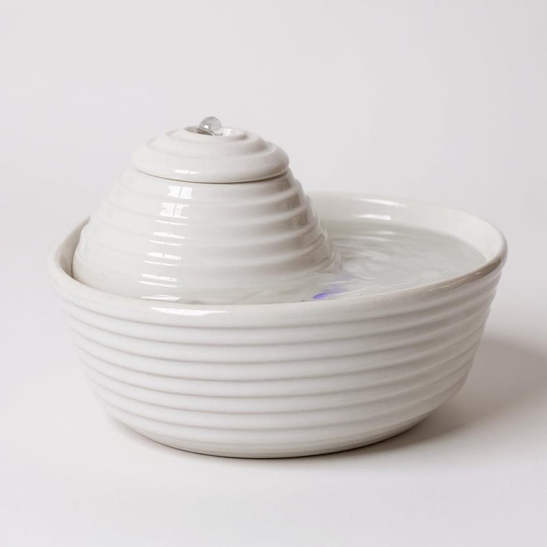 May include: A white ceramic pet fountain with a tiered, spiral design. The fountain has a bowl-shaped base and a central, cone-shaped feature with water gently flowing from the top. The water is clear, and the overall aesthetic is clean and modern.