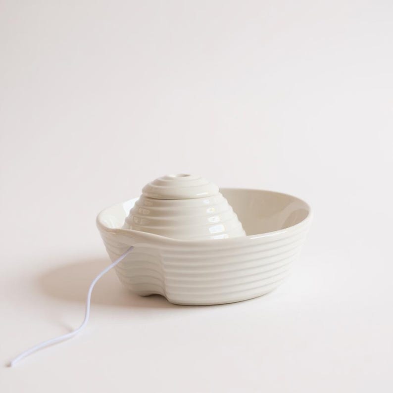 May include: A white ceramic citrus juicer. It features a ridged bowl and a central, cone-shaped reamer with concentric ridges. A white cord extends from the bowl. This kitchen tool is designed for juicing citrus fruits.