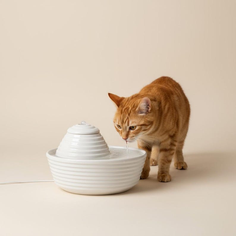May include: A white ceramic pet water fountain with a tiered design. A ginger tabby cat is drinking water from the fountain. The fountain has a round base and a small, decorative top. The cat has orange and white fur.