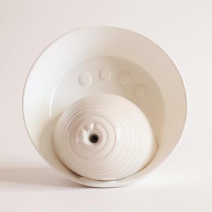May include: A white ceramic pet water fountain. The fountain has a bowl-shaped base and a central, domed fountain element. The fountain element has a textured, concentric circle design and a small spout. The bowl has a series of raised circles.