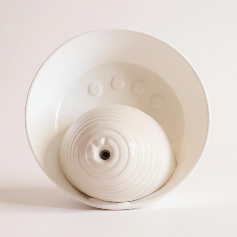 May include: A white ceramic pet water fountain. The fountain has a bowl-shaped base and a central, domed fountain element. The fountain element has a textured, concentric circle design and a small spout. The bowl has a series of raised circles.