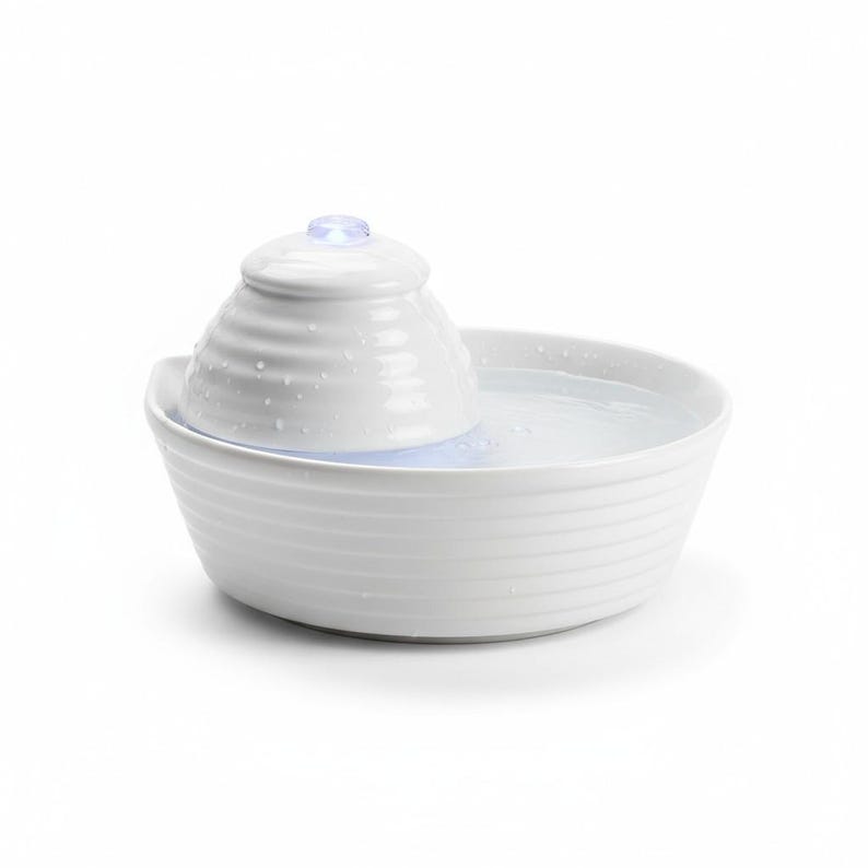 May include: A white ceramic pet water fountain with a tiered design. The fountain has a bowl-shaped base and a central, cone-shaped feature with a small, glowing light on top. Water is visible in the bowl.
