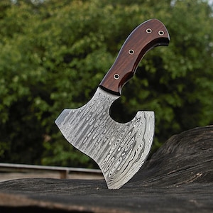 May include: A handcrafted axe with a dark wood handle and a patterned steel blade. The handle is curved and secured with rivets. The blade has a unique, textured design. The axe is displayed outdoors against a blurred green background.