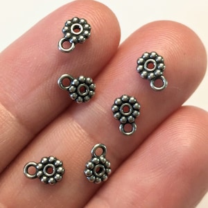 May include: Six silver-toned metal bead charms with a loop at the top. Each charm features a circle of beads surrounding a smaller circle.