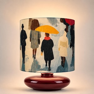 Może przedstawiać: Lampa stołowa z cylindrycznym kloszem ozdobionym ilustracją ludzi spacerujących w deszczu, w tym osoby trzymającej jaskrawożółty parasol. Lampa posiada gładką, błyszczącą, ciemnobordową ceramiczną podstawę o zaokrąglonym kształcie.
