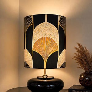 May include: A table lamp with a cylindrical shade featuring a black background and gold fan-shaped patterns. The lamp has a slender gold stem and a smooth, rounded black base. The light emits a warm glow from the top and bottom of the shade.
