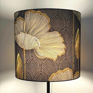 May include: A cylindrical lampshade with a gold and cream floral design set against a dark brown backdrop. The design includes a repeating fan-like pattern. The lampshade has a black trim. The lampshade would suit a living room or bedroom.