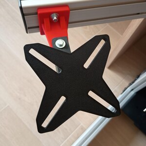 Peut inclure: Une plaque de montage métallique noire en forme de X fixée à un support en plastique rouge, lui-même attaché à un rail d'extrusion en aluminium argenté. La plaque comporte quatre fentes allongées pour le réglage et est fixée par des boulons.