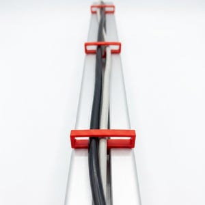 May include: A silver-colored metal cable management rail featuring three rectangular red plastic clips spaced along its length. Two electrical cables, one black and one white, are threaded through the clips and run along the center channel.