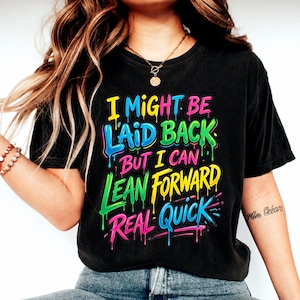Puede incluir: Camiseta negra con el texto "I Might Be Laid Back But I Can Lean Forward Real Quick" en una fuente colorida y goteante. La camiseta tiene un aspecto vintage.
