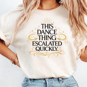 May include: A cream-coloured sweatshirt featuring the text "THIS DANCE THING ESCALATED QUICKLY" in black and gold. The design includes decorative swirls and stars. A casual garment.