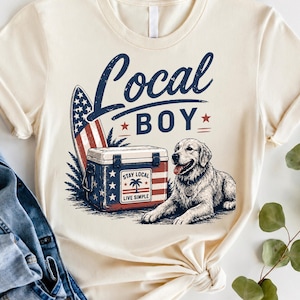 May include: A cream-colored t-shirt featuring a graphic of a golden retriever lying next to a cooler and a surfboard. The text reads Local BOY and STAY LOCAL LIVE SIMPLE. The design incorporates red, white, and blue colors with stars and stripes.