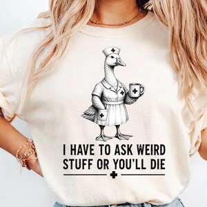May include: Cream-colored t-shirt featuring a black and white illustration of a goose dressed as a nurse, holding a mug. The text "I HAVE TO ASK WEIRD STUFF OR YOU'LL DIE" is printed below the goose.