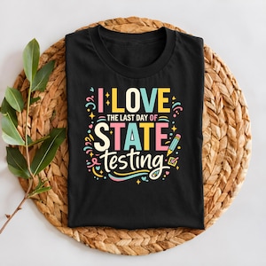 May include: Black t-shirt with the text "I LOVE THE LAST DAY OF STATE TESTING" in colorful, playful lettering. A pencil graphic is included. The shirt is folded on a woven, circular placemat.