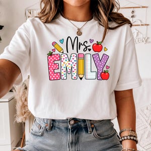 Puede incluir: Camiseta blanca con el texto "Mrs. Emily" en un diseño colorido y juguetón. El diseño incluye corazones, manzanas y lápices, con el nombre "Emily" en una fuente de lunares y líneas. Una camiseta con temática de profesor.