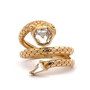 Può includere: Un anello in oro a forma di serpente con un motivo a scaglie testurizzato lungo la fascia. La testa del serpente è incastonata con una grande pietra chiara di forma pentagonale, mentre la coda presenta una pietra chiara più piccola a taglio marquise.