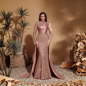 May include: A formal, floor-length gown in a dusty rose color. The dress features long sleeves, a high neckline, and a fitted bodice embellished with sequins. A draped satin overlay extends from the waist, creating a flowing train. The model is standing in front of a neutral background.