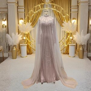 May include: A pale pink, floor-length gown with detailed beadwork and a high neckline. The dress has long sleeves and a flowing, sheer cape. The gown is displayed on a mannequin in a room with gold accents and decorative features, such as pampas grass.