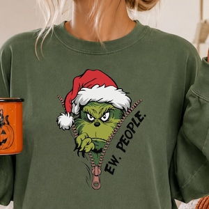 Pode incluir: Uma camisola de gola redonda verde-azeitona com um gráfico de uma personagem verde com um gorro de Pai Natal vermelho, a espreitar por trás de um fecho de correr com o texto "EW. PEOPLE." impresso a preto ao lado do fecho.