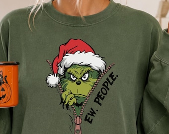 Peut inclure: Un sweat-shirt à col rond vert olive arborant un motif représentant un personnage vert portant un bonnet de Père Noël rouge, sortant d'une fermeture éclair avec le texte "EW. PEOPLE." imprimé en noir à côté de la fermeture.