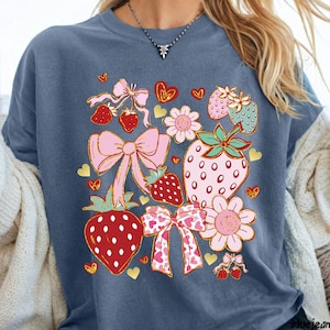 Pode incluir: Uma t-shirt azul com uma estampa gráfica de morangos vermelhos e cor-de-rosa, laços cor-de-rosa claros e pequenas flores. O design inclui formas de coração douradas espalhadas e uma mistura de elementos sólidos e estampados, criando uma ilustração.