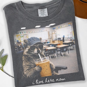 May include: Pepper-colored t-shirt featuring a raccoon wearing sunglasses and a denim cap, sipping from a coffee cup. The image on the shirt shows the raccoon in a classroom setting, with the text "i live here now".