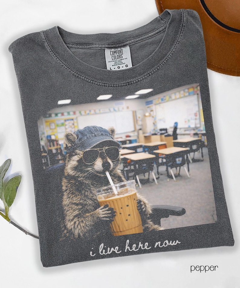 May include: Pepper-colored t-shirt featuring a raccoon wearing sunglasses and a denim cap, sipping from a coffee cup. The image on the shirt shows the raccoon in a classroom setting, with the text "i live here now".