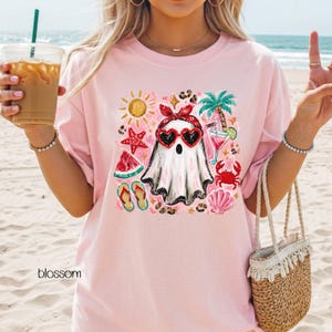 May include: Pink t-shirt featuring a ghost wearing heart-shaped sunglasses and a red bow, surrounded by beach-themed illustrations. The design includes a sun, watermelon, flip-flops, and a cocktail. The word "blossom" is printed at the bottom.