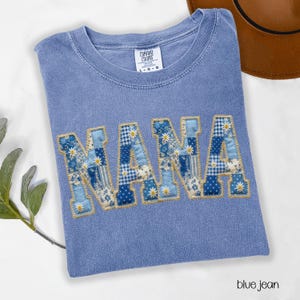 May include: A blue jean Comfort Colors t-shirt with the word "NANA" appliqued in a patchwork design. The letters feature various blue floral and patterned fabrics with daisy accents. The shirt is a light blue color.