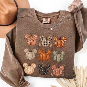 May include: A brown crewneck sweatshirt featuring a graphic print of nine pumpkins, each with mouse-ear shapes. The pumpkins display various patterns including leopard print, plaid, floral, polka dots, and solid colors. The label reads Comfort Colors.