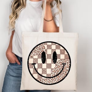 May include: Cream-colored tote bag featuring a large smiley face design. The smiley face is a combination of a checkered pattern and a leopard print pattern, with black eyes and a curved black smile. The bag has two long handles.