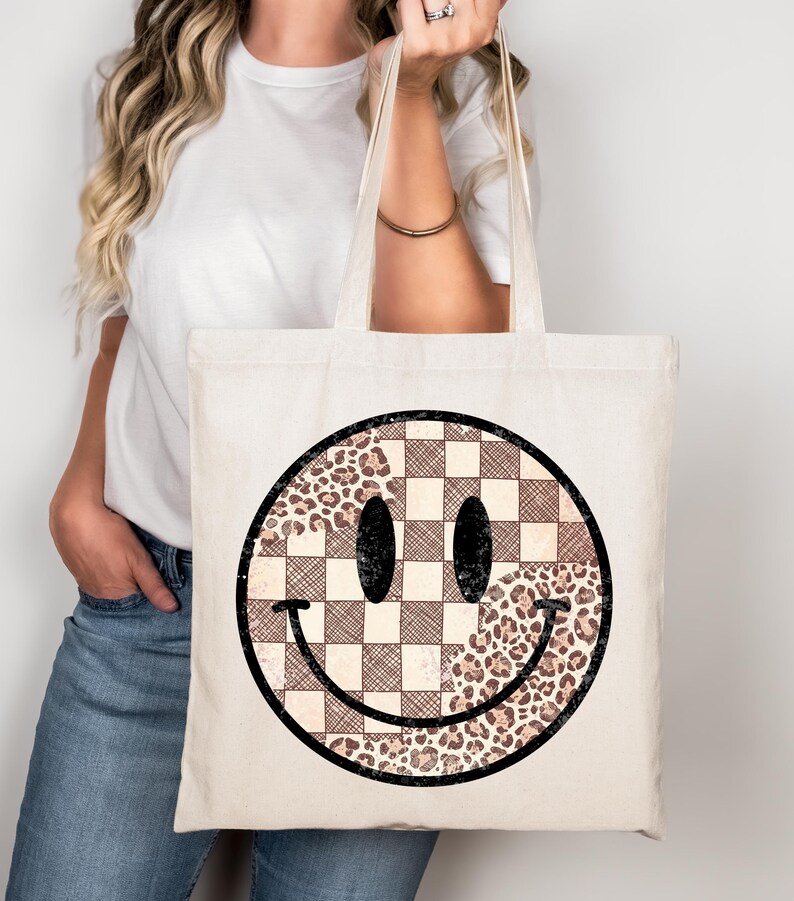 May include: Cream-colored tote bag featuring a large smiley face design. The smiley face is a combination of a checkered pattern and a leopard print pattern, with black eyes and a curved black smile. The bag has two long handles.