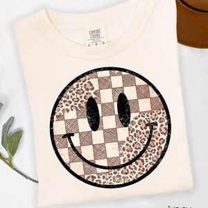 May include: Ivory-colored t-shirt featuring a large smiley face design. The smiley face is a combination of a checkered pattern and a leopard print pattern. The shirt has a crew neck and short sleeves. The brand label is visible on the upper left side.