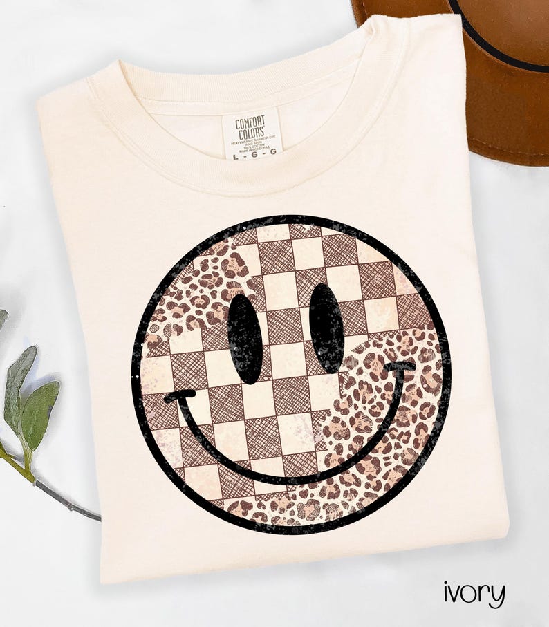 May include: Ivory-colored t-shirt featuring a large smiley face design. The smiley face is a combination of a checkered pattern and a leopard print pattern. The shirt has a crew neck and short sleeves. The brand label is visible on the upper left side.