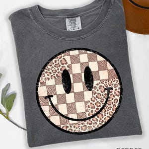 May include: Pepper-colored t-shirt featuring a large smiley face design. The smiley face is split into sections with a checkered pattern and a leopard print pattern. The shirt has a crew neck and short sleeves.