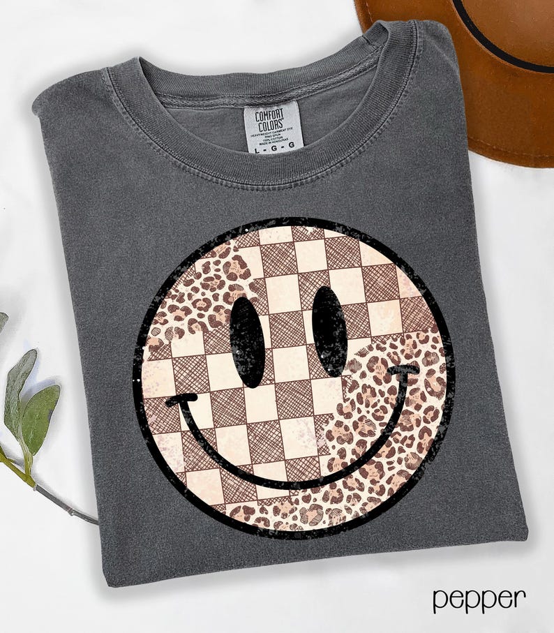 May include: Pepper-colored t-shirt featuring a large smiley face design. The smiley face is split into sections with a checkered pattern and a leopard print pattern. The shirt has a crew neck and short sleeves.
