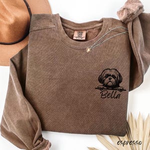 Puede incluir: Sudadera color espresso con cuello redondo y mangas largas. Un gr&aacute;fico negro de un perro Shih Tzu est&aacute; impreso en la parte delantera, con el nombre "Bella" debajo. Un collar plateado con un peque&ntilde;o colgante est&aacute; drapeado sobre la camisa.