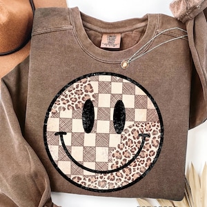 May include: Espresso-colored sweatshirt featuring a large smiley face design. The smiley face is split, with a checkered pattern on one side and a leopard print on the other. The word "espresso" is printed at the bottom.