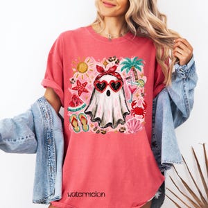 May include: A coral-colored t-shirt featuring a whimsical ghost design wearing heart-shaped sunglasses and a red bandana. The graphic includes beach-themed elements like a sun, palm tree, and watermelon. The word "watermelon" is printed at the bottom.