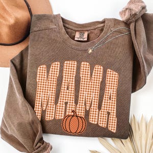 Pode incluir: Uma camisola castanha de gola redonda com a palavra MAMA em letras grandes de bloco aos quadrados cor de laranja e brancos. Abaixo do texto encontra-se um pequeno gr&aacute;fico de uma ab&oacute;bora cor de laranja com pintas. Etiqueta Comfort Colors.