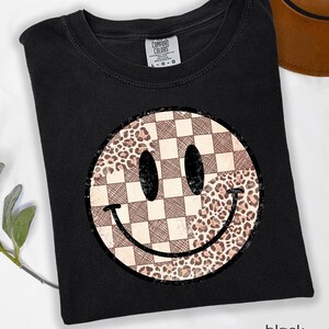May include: Black t-shirt featuring a smiley face design. The smiley face has a checkered pattern on one side and a leopard print on the other. The shirt is labeled "Comfort Colors" and the word "black" is printed at the bottom.
