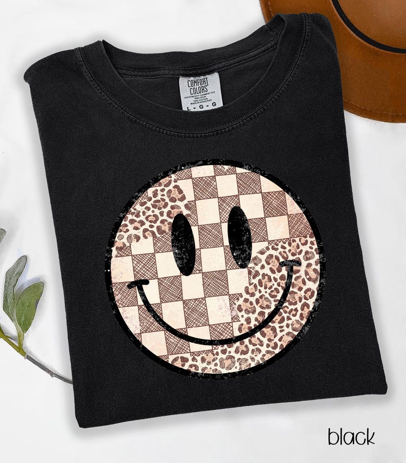 May include: Black t-shirt featuring a smiley face design. The smiley face has a checkered pattern on one side and a leopard print on the other. The shirt is labeled "Comfort Colors" and the word "black" is printed at the bottom.