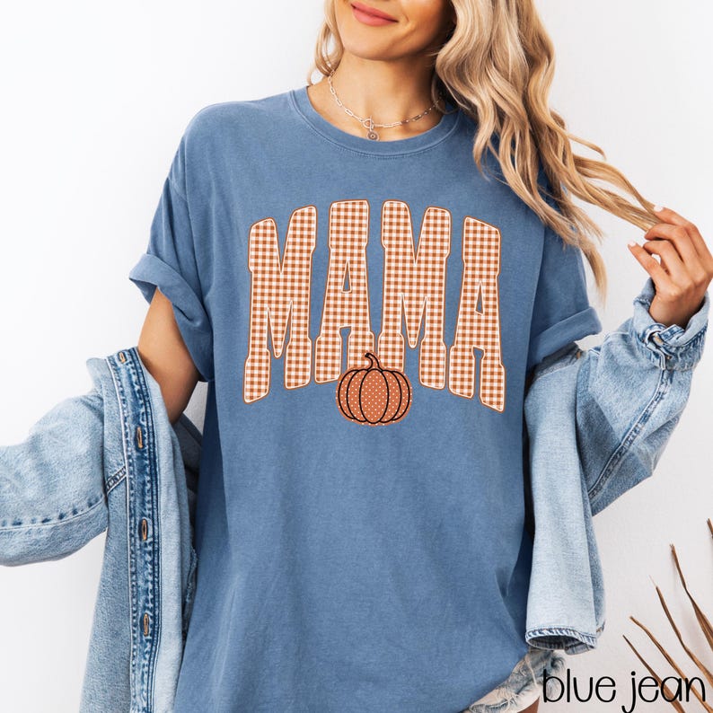 Pode incluir: Uma t-shirt azul de manga curta com a palavra MAMA numa fonte grande de xadrez vichy laranja e branco. Abaixo do texto, h&aacute; um pequeno gr&aacute;fico de uma ab&oacute;bora laranja com bolinhas brancas. A t-shirt tem gola redonda e corte descontra&iacute;do.