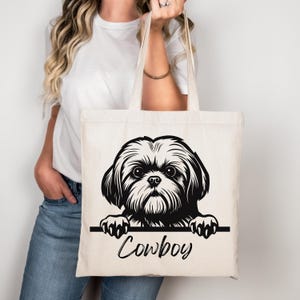 Puede incluir: Un bolso tote de color crema con una ilustraci&oacute;n en blanco y negro de un perro Shih Tzu. Se ve la cabeza y las patas del perro, con el nombre "Cowboy" escrito en cursiva debajo. El bolso lo sostiene una persona.
