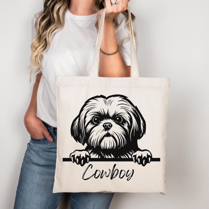 Puede incluir: Un bolso tote de color crema con una ilustraci&oacute;n en blanco y negro de un perro Shih Tzu. Se ve la cabeza y las patas del perro, con el nombre "Cowboy" escrito en cursiva debajo. El bolso lo sostiene una persona.