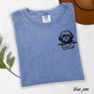 Puede incluir: Camiseta azul vaquero Comfort Colors con un gr&aacute;fico negro de un perro Shih Tzu y el nombre "Bella". La camiseta tiene cuello redondo y mangas cortas. Un sombrero marr&oacute;n es visible en el fondo.