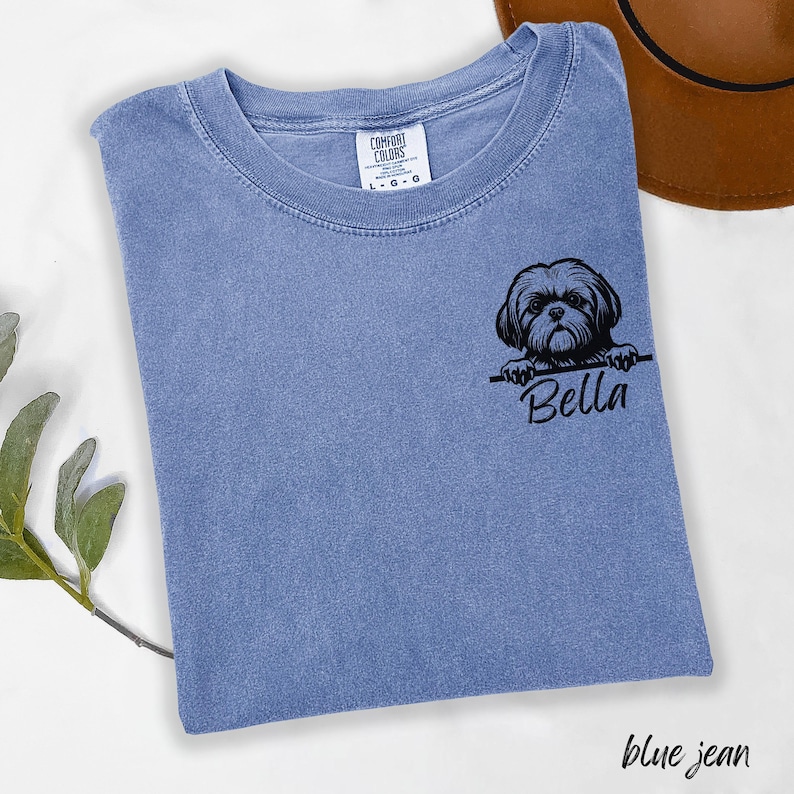 Puede incluir: Camiseta azul vaquero Comfort Colors con un gr&aacute;fico negro de un perro Shih Tzu y el nombre "Bella". La camiseta tiene cuello redondo y mangas cortas. Un sombrero marr&oacute;n es visible en el fondo.