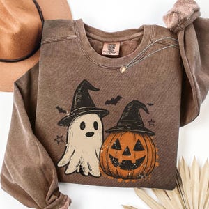 Pode incluir: Uma camisola castanha de gola redonda com uma impressão gráfica de um fantasma e uma abóbora, ambos a usar chapéus de bruxa pretos. O design inclui pequenos morcegos e estrelas. A etiqueta interior apresenta o texto Comfort Colors.
