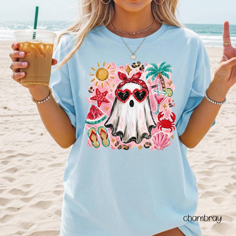 May include: Light blue t-shirt with a colorful graphic of a ghost wearing heart-shaped sunglasses, a red headband, and surrounded by beach-themed illustrations. The word "chambray" is printed on the bottom right.
