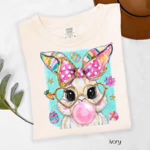 Peut inclure: T-shirt ivoire avec un motif de lapin de P&acirc;ques fantaisiste. Le lapin porte des lunettes en forme de c&oelig;ur et un n&oelig;ud rose, soufflant une bulle de chewing-gum. Des &oelig;ufs de P&acirc;ques et des fleurs color&eacute;s entourent le lapin sur un fond turquoise.